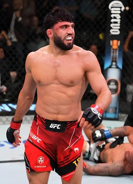 The image shows a UFC fighter celebrating a victory inside the octagon, wearing red UFC shorts with "UFC" visible on the waistband, while another fighter lies on the mat behind him; this victory scene relates to the accompanying post about betting on UFC fights, including Arman Tsarukyan vs. Mauricio Ruffy and Joshua Van vs. Alexandre Pantoja.
