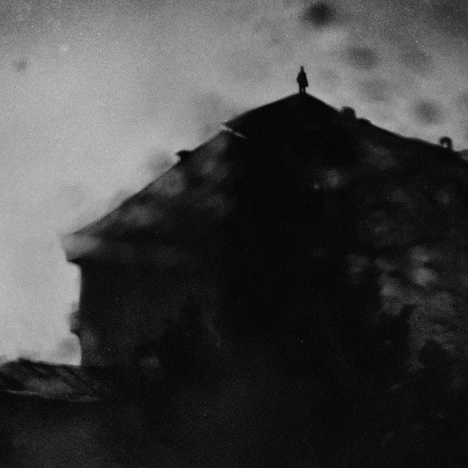 A moody black and white photograph showing a lone figure silhouetted atop a rooftop against a hazy, overcast sky, creating an ominous atmosphere.