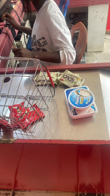 A shop counter displays small red packets in a wire basket, a FanIce container, and some money, with a person seated behind a glass partition holding cash.