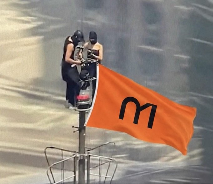 Two masked individuals sit atop a tall pole or mast, unfurling a large orange flag with a black 'M' logo.