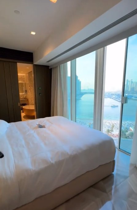 A luxurious high-rise hotel room features a neatly made king-size bed with a small towel animal, floor-to-ceiling windows revealing a marina view, and an open bathroom doorway—evoking a "good morning" from a beautiful travel destination. The opulent setting complements the post's upbeat GM greeting, symbolizing an aspirational lifestyle of waking up in paradise.