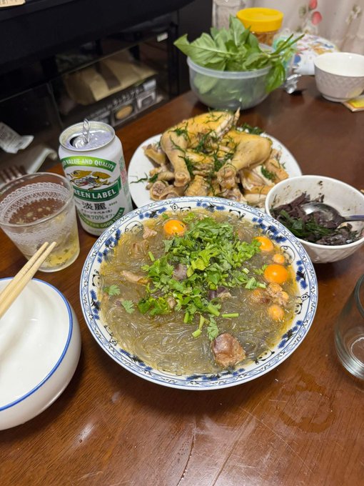 A home-cooked meal on a wooden table featuring a bowl of glass noodles with vegetables and quail eggs, chicken pieces, fresh herbs, and a can of Suntory Premium Malt's beer.