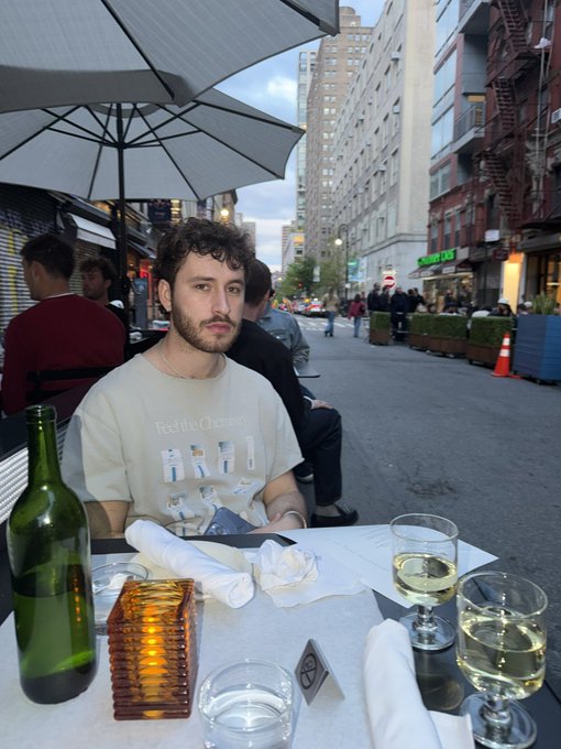 The image shows a man sitting at an outdoor table with wine glasses, including one filled with white wine. There are also two other people present in the scene, but they do not appear to be engaged in conversation or dining activities. The setting appears to be a restaurant patio, and there is a bottle of wine on the table as well.