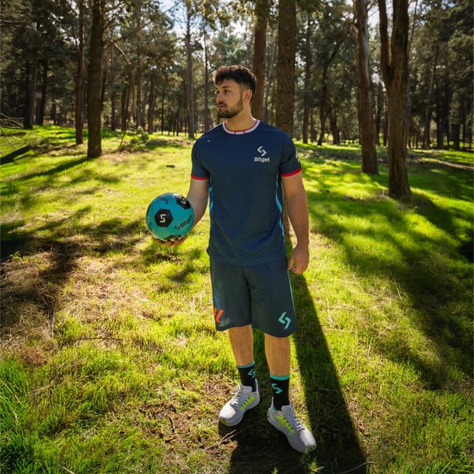 A man stands in a sunlit forest wearing a navy athletic kit with "Bitget" branding, holding a teal and black soccer ball, ready for an outdoor match. The "Bitget" sponsorship logo and sportswear connect to the "All set for match day?" post, highlighting preparedness for a game in a scenic natural setting.