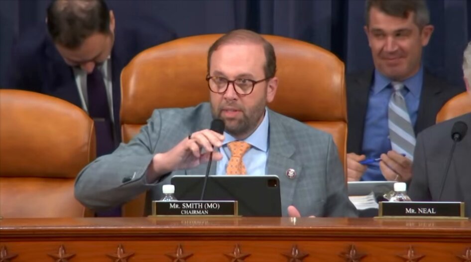 The image shows a group of people sitting around a table, with one man wearing glasses and holding a laptop. The scene appears to be taking place at a hearing or meeting related to Bitcoin tax reform. The room has a gray color scheme, and there are chairs placed around the table where the attendees are seated. One person is seen using a cell phone, possibly checking messages or looking up information about the ongoing discussion on Bitcoin tax reform.