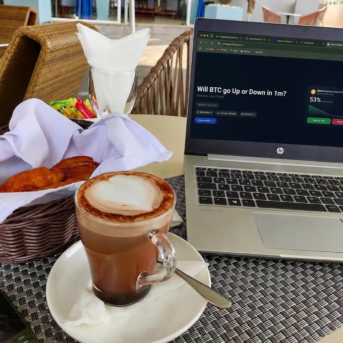 A laptop displays a Polymarket prediction page titled 'Will BTC go Up or Down in 1m?' with 53% chance shown, next to a cappuccino and pastries on a cafe table.