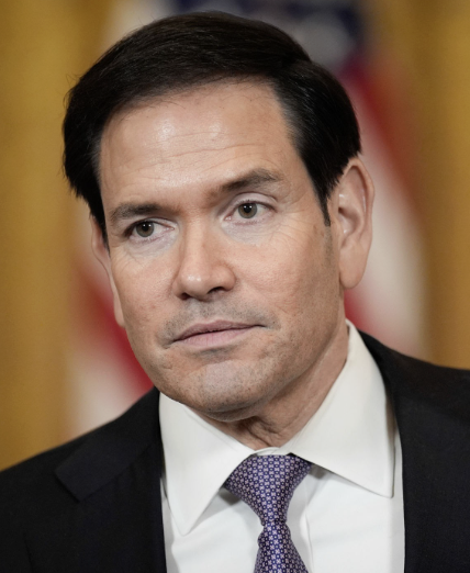 Secretary of State Marco Rubio, wearing a dark suit and purple patterned tie, listens intently during a press briefing at the State Department. The image accompanies news that Rubio has informed allies the U.S. is shifting from military strikes to economic warfare against Iran, including the seizure of nearly $1 billion in cryptocurrency.