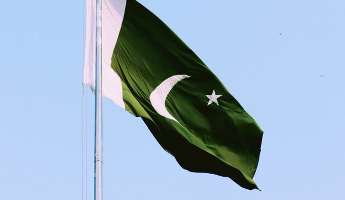A photograph of the Pakistani national flag—green with a white crescent and star, and a white vertical stripe—waving on a flagpole against a clear blue sky.