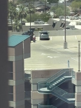 View from an upper floor of a parking lot with cars parked, palm trees in the distance, and a teal-roofed building with stairs in the foreground.