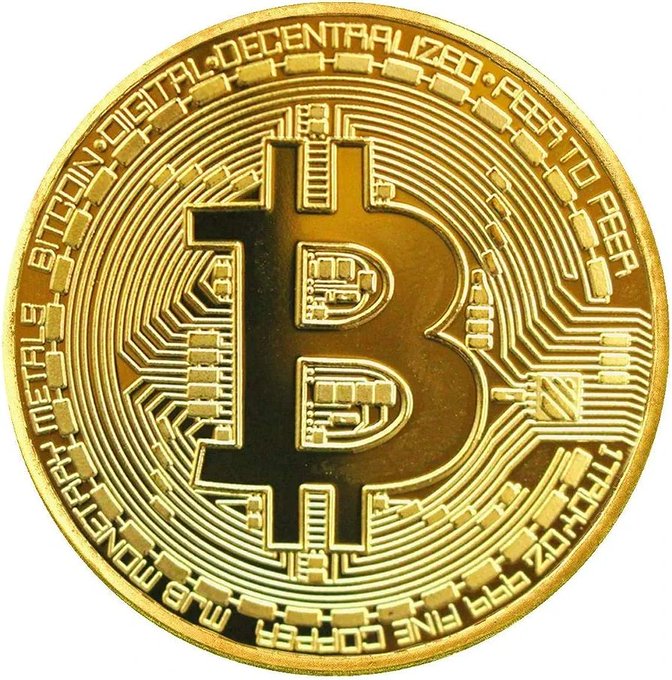 A physical gold-colored commemorative Bitcoin coin featuring the 'B' logo with text 'BITCOIN DIGITAL DECENTRALIZED PEER TO PEER' and 'VIRES IN NUMERIS FINE COPPER' around the edge.