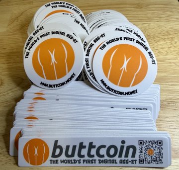 Two stacks of round and rectangular "Buttcoin" stickers, branded as "The World's First Digital Ass-Set," are displayed on a wooden surface, illustrating the promotional merchandise for the giveaway. The stickers prominently feature the Buttcoin logo and website (www.buttcoin.org) along with a QR code for the digital currency.