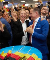 A man in a blue suit celebrates joyfully, embracing another person in a crowd while surrounded by colorful balloons and clapping supporters.
