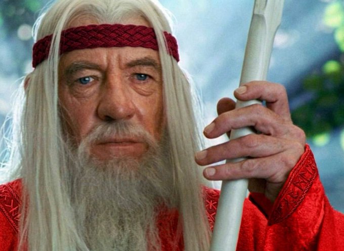 An elderly man with long white hair and beard, wearing a red braided headband and red robe, grips a silver staff with a serious expression. The image, appearing to be from a film featuring a wise wizard-like character, aligns with the post's hype for an exciting "$GME" video being released tonight.