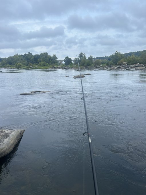 A fishing rod extends over a calm, overcast river dotted with rocks, with a person wading in the distance near a bridge. The moody, gray sky and still water set the perfect stage for "bad day to be a fish."