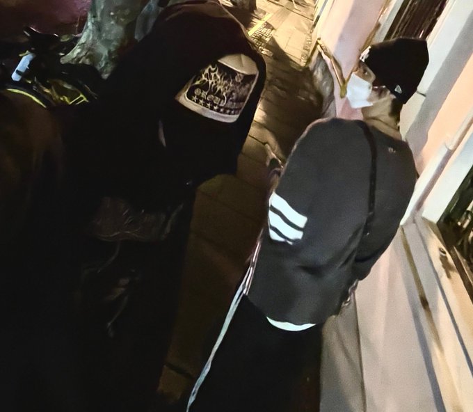 A dimly lit image of a person wearing a black jacket with white stripes on the sleeve, a black beanie, and a white face mask, viewed from behind near a car.