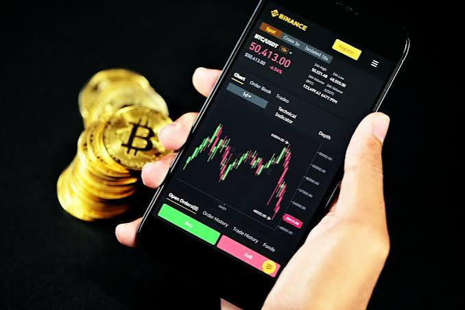 A hand holds a smartphone displaying the Binance trading app for BTC/USDT at $50,413.00, with gold Bitcoin tokens stacked next to the phone.