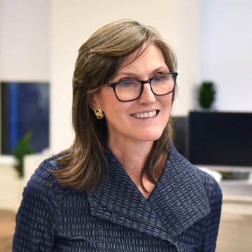 A portrait of a smiling woman with shoulder-length hair wearing black-rimmed glasses and a patterned navy blazer.