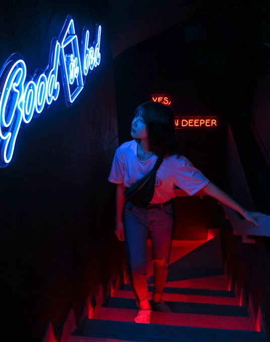 The image features a woman standing at the bottom of stairs, looking up towards a neon sign that says "Good Night Fam Leaving all today's thoughts here for now for now. Rest. Sleep. Dream." The woman appears to be wearing a white shirt and blue jeans. She is holding onto the handrail while standing on the stairs.