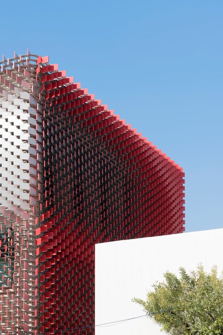 A modern architectural facade with a distinctive red lattice or pixelated screen structure against a clear blue sky, with a tree visible at the bottom right.