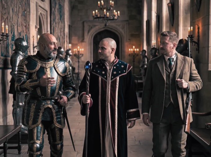 Three men stand in a castle hallway; one wears ornate armor, the center man wears a dark robe with star patterns and holds a staff, and the right man wears a brown suit.