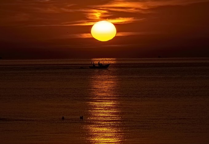 A serene sunset over the ocean with a silhouetted boat on the water, the bright sun reflecting a golden path across the sea.