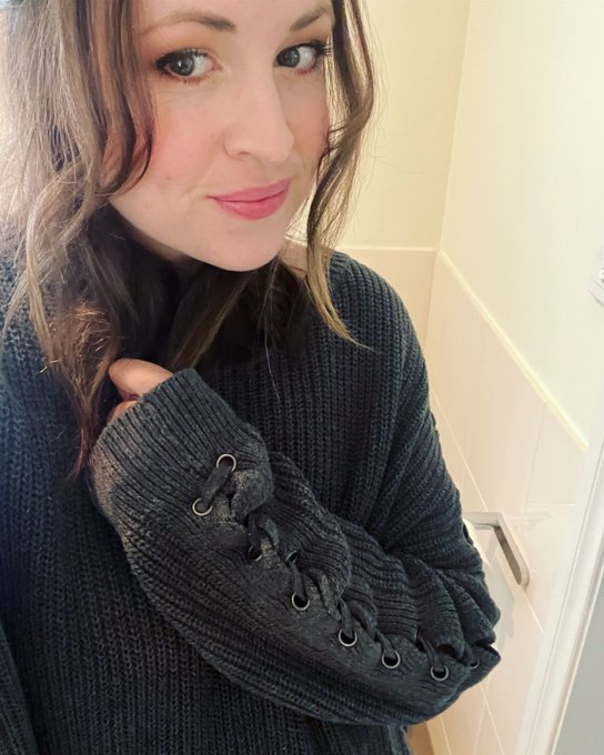 A selfie of a smiling woman with long brown hair wearing a dark gray knit sweater featuring lace-up sleeve detail, taken in what appears to be a bathroom.