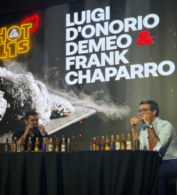Two men sit at a table lined with hot sauce bottles, simultaneously biting into chicken wings at the "Hot Ones" show, with the backdrop reading "LUIGI D'ONORIO DEMEO & FRANK CHAPARRO." The image captures their first wing attempt out of ten, matching the post's countdown progress.