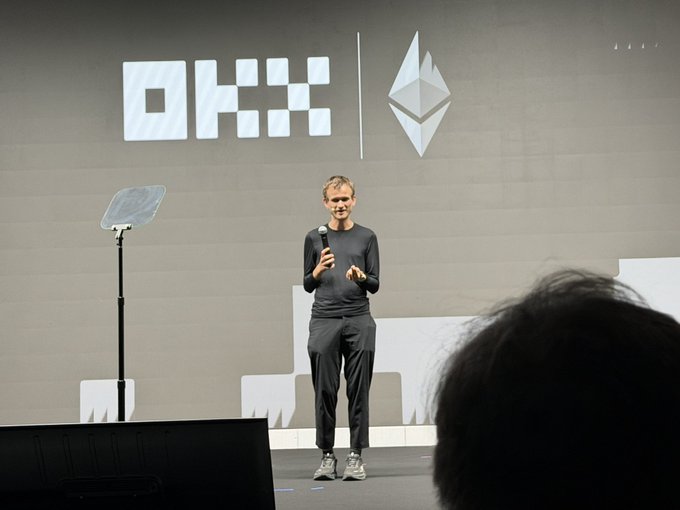 A man stands on stage presenting in front of a large OKX logo and Ethereum symbol backdrop.