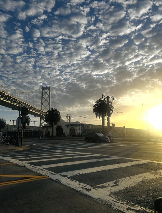 A scenic sunset view of the San Francisco-Oakland Bay Bridge with palm trees silhouetted against a dramatic cloud-filled sky, with a crosswalk and street intersection in the foreground.