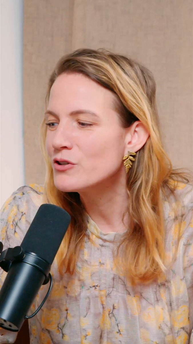 The image features a woman wearing a floral print dress, sitting at a microphone while talking into it. The background of the image is a plain white wall. There are no other objects or people visible in the image.
