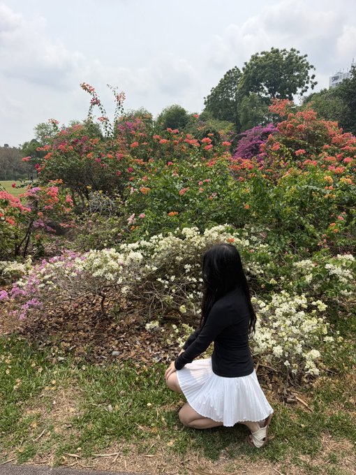 A woman with long dark hair, wearing a black top and white skirt, kneels on the grass to admire a lush garden full of blooming pink, red, and white flowers.