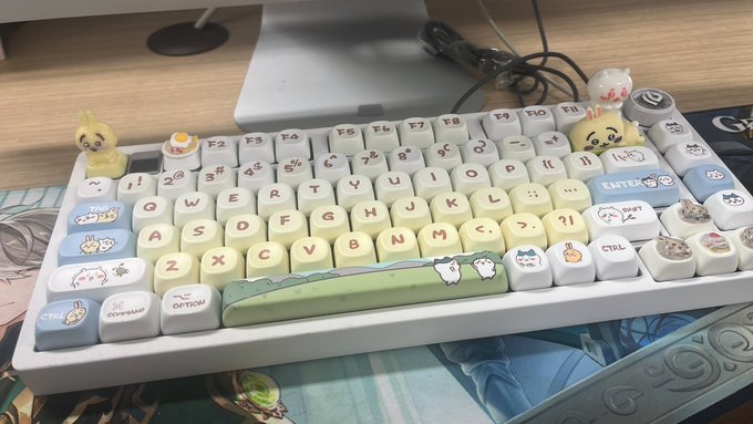 A 65% mechanical keyboard decorated with cute cat-themed keycaps and small figurines (including a chick and what appears to be a Pompompurin character) sits on a desk mat, with visible keys like "SHIFT," "CTRL," "OPTION," "COMMAND," and "ENTER." The image relates to the post by illustrating how distinctive, eye-catching details—like the whimsical animal designs on the keyboard—draw attention, reinforcing the message that what captures notice becomes leverage.