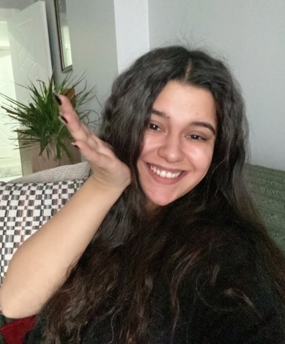 A smiling young woman with long, curly dark hair waves warmly at the camera in a cozy living room setting. The cheerful, welcoming pose perfectly matches her friendly reintroduction, inviting others to share their artwork in the Tezos community after a long absence.