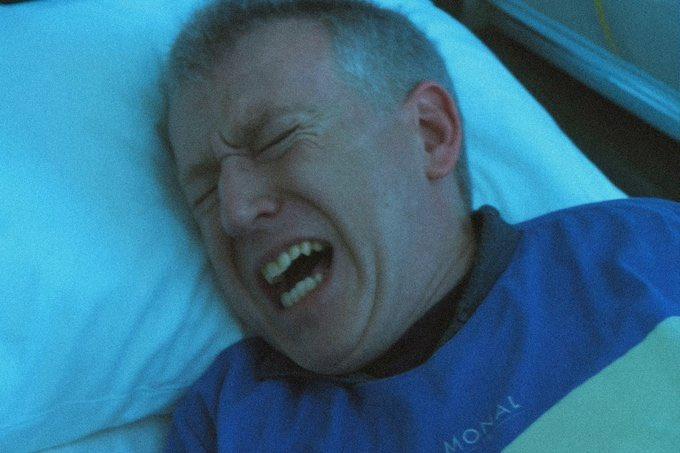 A close-up of a man lying on a white pillow, eyes shut tight and mouth open in an expression of pain or distress, wearing a blue shirt with 'MORAL' partially visible.
