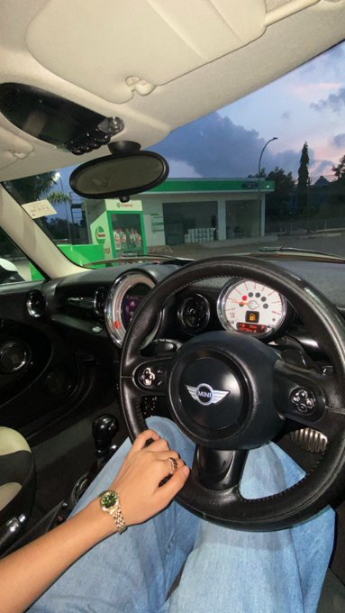 The image shows a first-person view from the driver's seat of a Mini Cooper, with a hand wearing a watch resting on the steering wheel while parked at a green-themed BP gas station at dusk, perfectly matching the post's playful reference to "touching the green MiniC instead of grass."