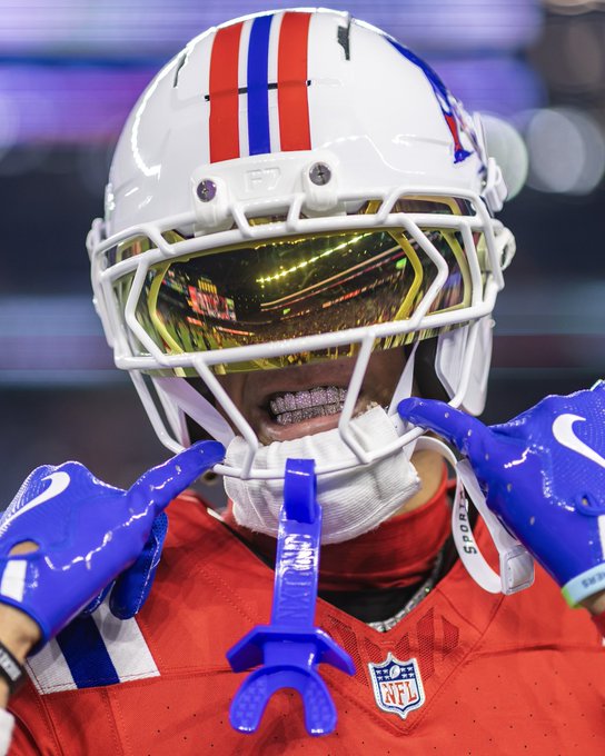 A close-up of a football player wearing a white helmet with red and blue stripes and a gold-tinted visor, showing their teeth and braces.