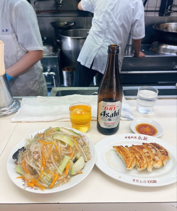 A meal at what appears to be a Chinese restaurant showing a plate of stir-fried glass noodles with vegetables, gyoza dumplings on a '永和' (Yonghe) branded plate, a glass of beer, and a bottle of Asahi Super Dry beer, with chefs working in the kitchen background.