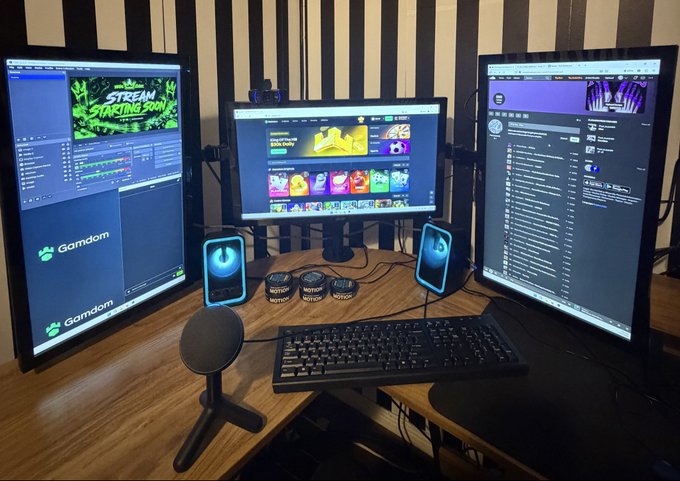 A streaming/gaming setup featuring three monitors (one showing 'STREAM STARTING SOON' graphics, others displaying Gamdom and other apps), a microphone, keyboard, and illuminated phone holders on a wooden desk