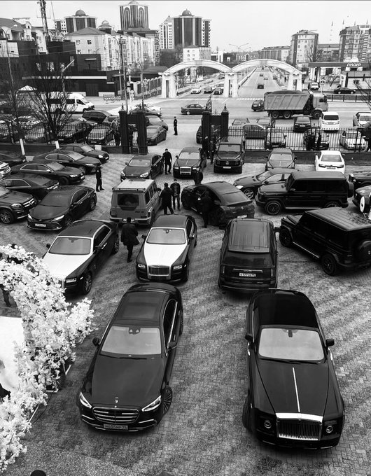 A black-and-white aerial view shows a fleet of luxury vehicles—including several black Mercedes-Benz, a Rolls-Royce, and a Bentley—lined up in a paved courtyard beside a stone archway entrance, with people in dark suits milling about and a cityscape and "DRIVING" parking building in the background, fitting the post's "Crypto bros meet up in 2030" caption.