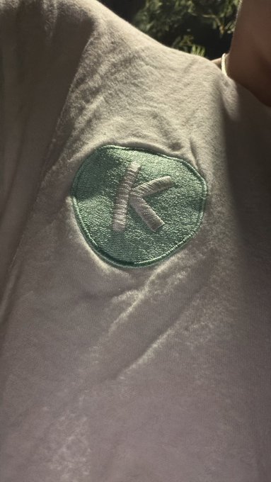 A close-up of a cream-colored tee features a circular green embroidered logo with a stylized "K" in the center. The image complements the caption by showcasing the "KAS" branded shirt being worn during an evening walk.
