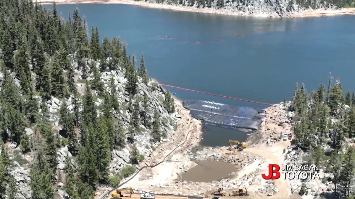 An aerial view shows a reservoir undergoing construction or repair work, with construction equipment and vehicles on the rocky shoreline near a large dark tarp, branded with 'Jim Bagan Toyota' in the corner.