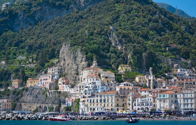 A photograph of a coastal Mediterranean town with colorful buildings clustered along the shoreline at the base of a steep, green mountain, with small boats in the blue water.
