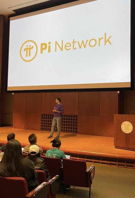 A speaker presents at an internal Pi Network meeting, with the "Pi Network" logo and name displayed prominently on a large projection screen behind him, as the team discusses ecosystem building efforts beyond trading to include applications, goods, and services.