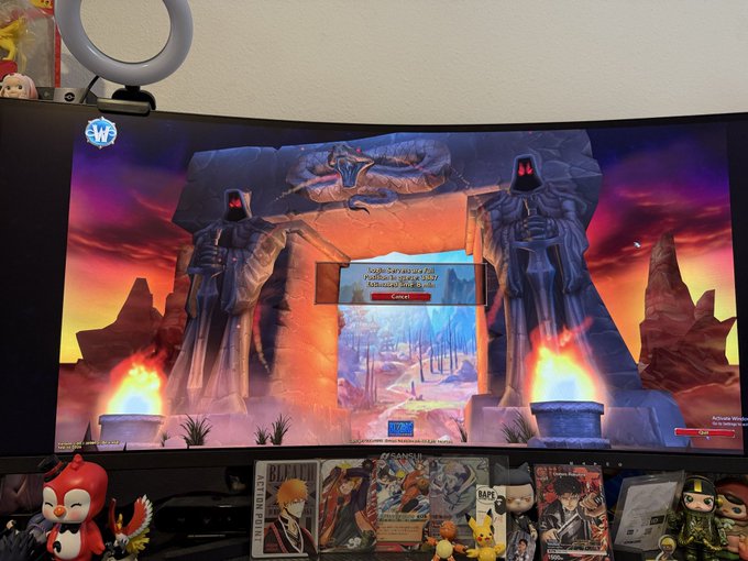 A curved monitor displays a fantasy video game with statues and a fiery portal, surrounded by anime figurines and manga volumes.