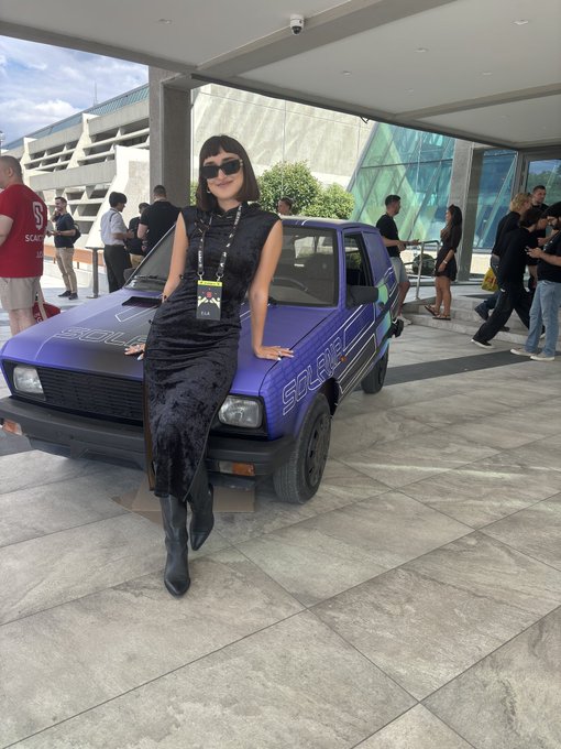 A woman in a black dress and sunglasses posing in front of a purple Solana-branded car at an outdoor event venue.