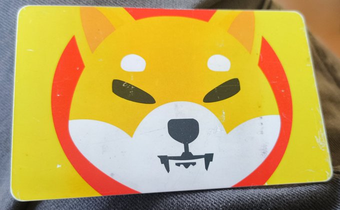 A yellow and red card or sticker with a stylized Shiba Inu dog face design, resting on a textured surface.