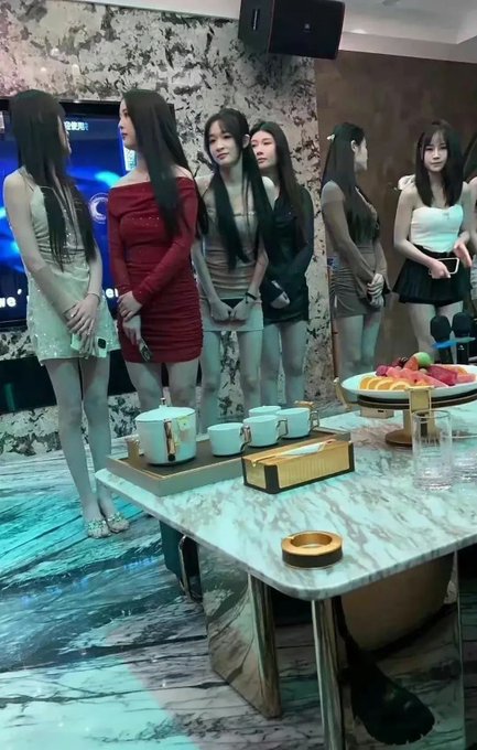 A group of young women in dresses stand together in what appears to be a karaoke room with marble walls, near a table with a fruit platter and tea set.