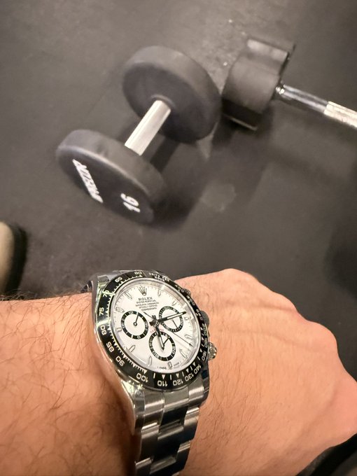 A person's wrist displays a Rolex Cosmograph Daytona watch with a white dial and black bezel, photographed in a gym setting with a 15 lb Life Fitness dumbbell on the floor.