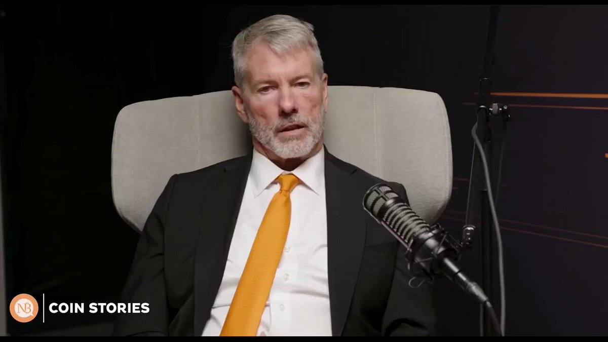The image shows an older man wearing a suit and tie sitting behind a microphone, likely participating in a podcast or interview. He has gray hair and appears to be speaking into the microphone. The background features a black wall with white text that reads "coin stories".