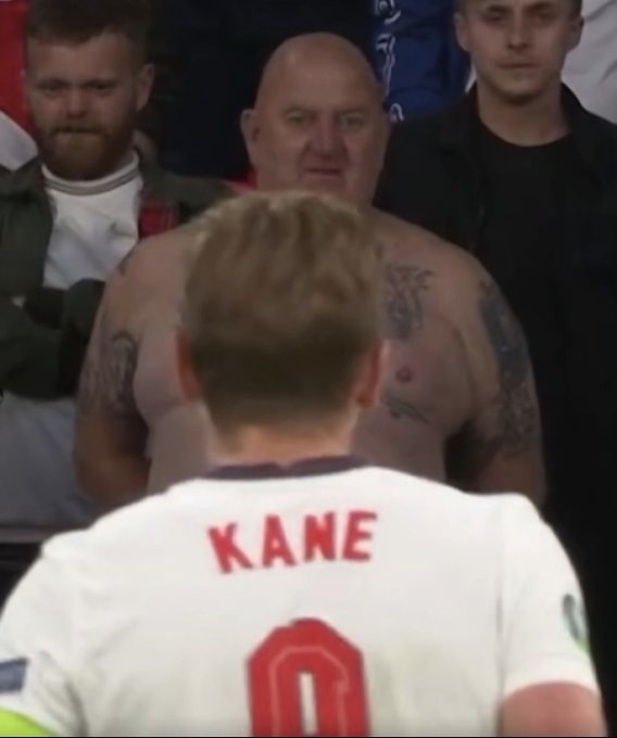 The image shows a man wearing a white jersey with red lettering, possibly representing a soccer team or player named Kane. The man has tattoos on his chest and appears to be bald. He is standing next to another person who is also wearing a white jersey but without any tattoos. They are both looking at the camera. In the background, there are other people present in the scene.